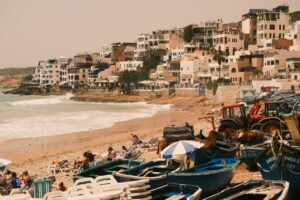 Scenic view of Taghazout beach with colorful boats, sunbathers, and adjacent hillside buildings.